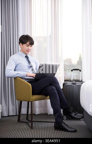 Ein junger Geschäftsmann arbeitet in einem Hotelzimmer an seinem Laptop und blickt aus dem Fenster auf die Skyline der Stadt. Stockfoto