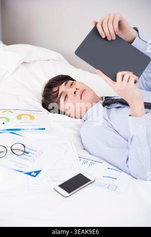 Ein junger Geschäftsmann liegt im Bett und liest auf seinem Tablet, inmitten eines verstreuten Chaos von Papieren und Diagrammen. Stockfoto