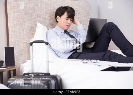 Ein junger Geschäftsmann sitzt im Bett und arbeitet an seinem Laptop, während er sich auf eine Reise vorbereitet. Stockfoto