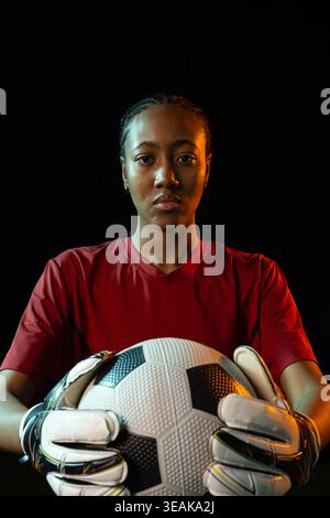 Afroamerikanische Hüterin hält B und W Fußball auf der Brust und trägt rote Jerseyhandschuhe im Studio. Torwart, Porträt, Studio, dramatisch, Beleuchtung, Stockfoto