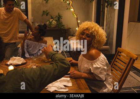 Person in grüner Jacke gießt Wasser, Frau lächelt an Tisch, animierte Glasfüllung zum Essen. Abendessen, Treffen, Freunde, lässig, gemütlich, warme Terrasse Stockfoto