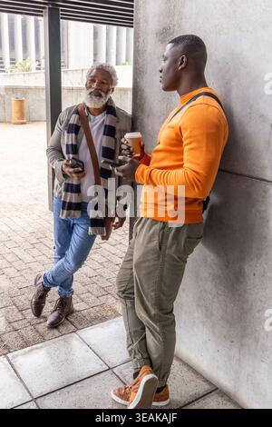 Afroamerikanische Männer, darunter Senioren, die sich im Innenhof mit Smartphones und Kaffee unterhalten. Urban, plaza, Outdoor, Beton, Telefon, Conversati Stockfoto