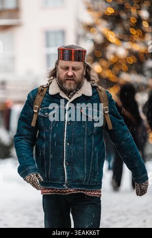 Tallinn, Estland - 03. Januar 2025: Blick auf einen Mann mit traditionellem Hut, der durch einen schneebedeckten Tallinner Weihnachtsmarkt geht, die festlichen Lichter verwischen Stockfoto