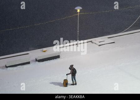 Tallinn, Estland - 03. Januar 2025: Blick auf einen einsamen Reisenden mit gelbem Koffer, der auf einer schneebedeckten Straße in der Nähe des Ufers unter dem Sof steht Stockfoto