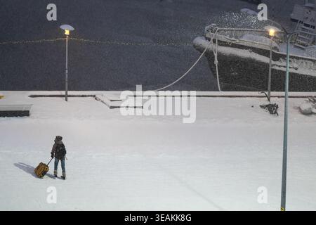 Tallinn, Estland - 03. Januar 2025: Blick auf eine Einzelfigur, die einen Koffer über einen schneebedeckten Pier zieht, die Silhouette eines Schiffes im Hintergrund Stockfoto