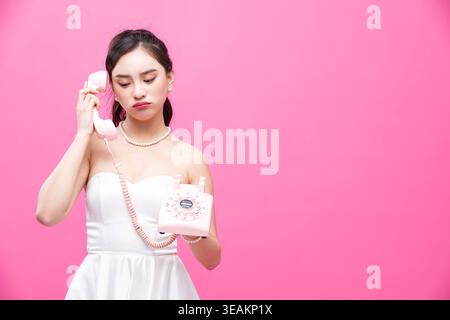 Eine Frau in einem weißen Kleid hält ein rosafarbenes Drehtelefon und blickt mit einem nachdenklichen Ausdruck nach unten. Stockfoto