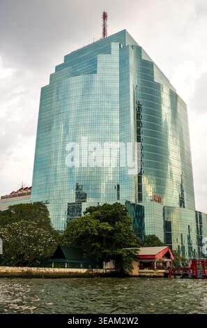 Moderner Glashochhaus, der sich über umliegenden Gebäuden und tropischen Bäumen im Stadtzentrum von Bangkok erhebt und den blauen Himmel mit Wolken reflektiert. Stockfoto