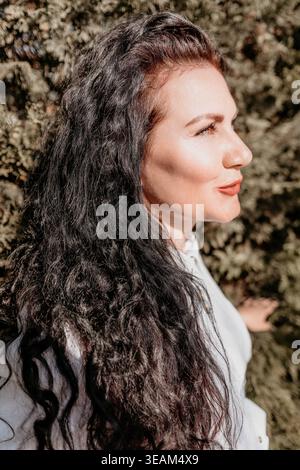 Frau Gartenporträt lächelnde Erwachsene Frau mit langen welligen Haaren steht draußen zwischen grünem Laub in weichem Sonnenlicht Stockfoto