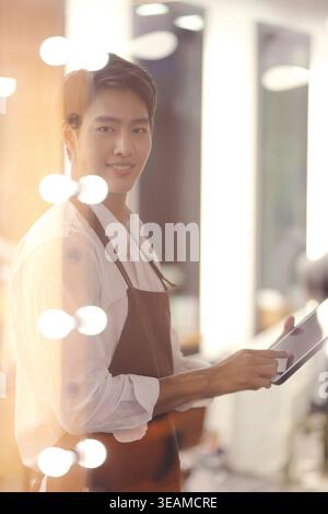Ein junger asiatischer Mann in einem Friseurladen lächelt, während er eine Tafel hält und über den Haarschnitt seines Kunden nachdenkt. Stockfoto