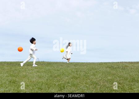 Zwei Kinder laufen zusammen auf einem grünen Grasfeld und spielen mit bunten Ballons. Stockfoto