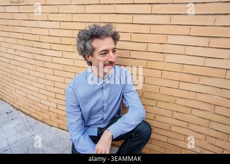Mann mittleren Alters mit grauen lockigen Haaren und Bart, trägt ein blau gestreiftes Hemd, hockt sich draußen an einer warmen Ziegelwand Stockfoto