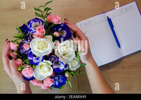 Weibliche Hände halten eine Rosenblume. Notizbuch, Tagebuch mit einem Stift auf einem Holztisch in der Nähe eines Straußes aus mehrfarbigen Rosen. Stockfoto
