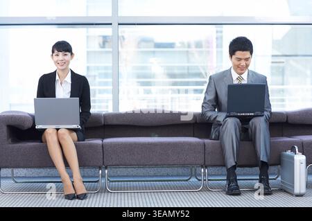 Zwei Geschäftsleute sitzen in einem modernen Büro auf einer Couch und arbeiten beide an ihren Laptops. Stockfoto