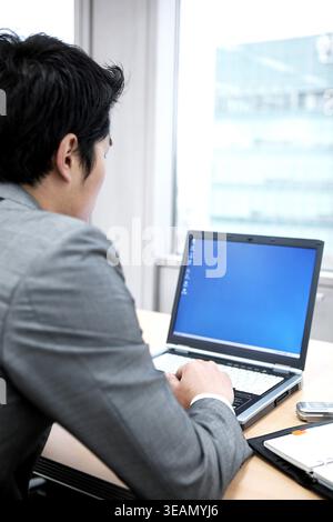 Ein Mann in einem grauen Anzug sitzt an einem Schreibtisch vor einem Laptop mit leerem Bildschirm. Stockfoto