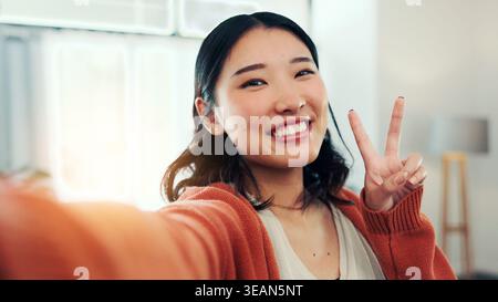 Glückliche Frau, Porträt und Selfie mit Friedenszeichen zu Hause, Profilbild oder Erinnerung für soziale Medien. Influencer, Online- und asiatische Person mit Lächeln Stockfoto