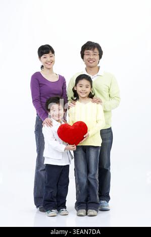 Eine glückliche vierköpfige Familie mit zwei Kindern und ihren Eltern, die zusammen steht und in die Kamera lächelt und ein herzförmiges Kissen hält. Stockfoto