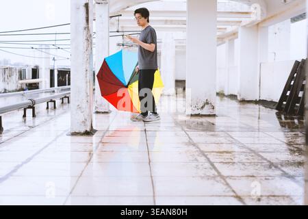Junger Mann mit buntem Regenschirm, der auf einer nassen, gekachelten Plattform unter weißen Säulen läuft, regnerischer Tag Stockfoto