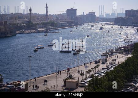 Blick aus der Vogelperspektive auf traditionelle Abra-Boote, die den Dubai Creek überqueren, mit Menschen, die entlang der Uferpromenade und historischen Gebäuden im Hintergrund spazieren Stockfoto