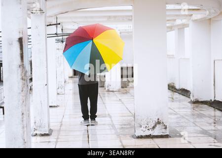 Person mit farbenfrohem Regenbogenschirm unter weißem Betongestell an regnerischen Tagen, minimalistisch Stockfoto