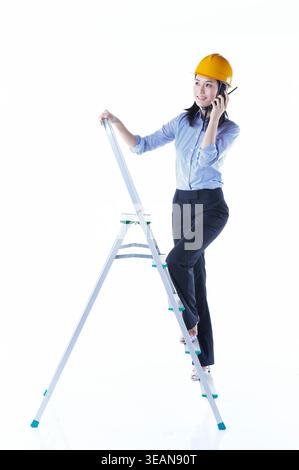 Eine Frau mit Schutzhelm steht auf einer Leiter, während sie ein Telefon an ihr Ohr hält. Stockfoto