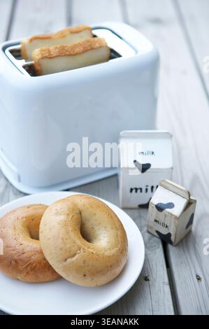 Auf einem Holztisch steht ein weißer Toaster, im Hintergrund wird Brot getoastet, im Vordergrund befindet sich ein Teller Bagels und ein Milchkarton. Stockfoto