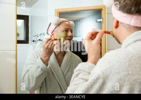 Reifer Mann, der grüne Ton Gesichtsmaske mit Pinsel aufträgt, lustiges rosafarbenes Stirnband und Bademantel trägt, authentische männliche Schönheit Routine und Hautpflege Stockfoto
