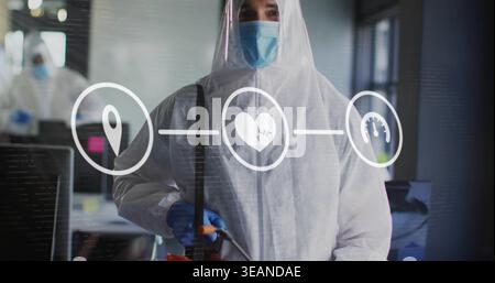Hält Desinfektionsstab, Person trägt weiße PSA, blaue Maske, die Büromonitore sprüht, HUD-Symbole. PSA, Handschuhe, Overalls, Gefahrgut, Hygiene, Teamwork Stockfoto