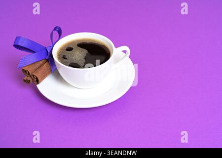 Ein hochwertiges Nahfoto einer weißen Tasse mit schwarzem Kaffee auf einer Untertasse, begleitet von Zimtstangen, die mit einem lila Band gebunden sind Stockfoto