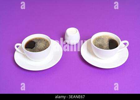 Zwei Tassen schwarzen Kaffee mit Milchkännchen auf lila Hintergrund Stockfoto