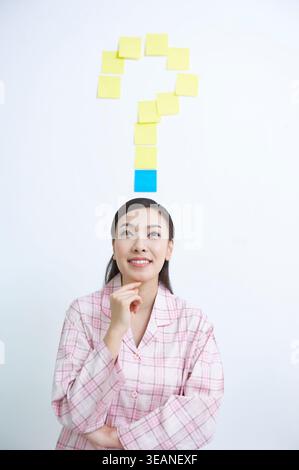 Eine Frau in einem rosa-weißen karierten Pyjama-Set schaut auf ein Fragezeichen aus Haftnotizen. Stockfoto