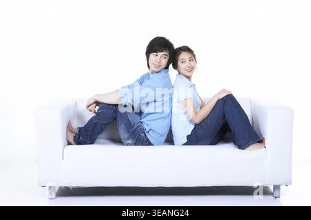 Ein junges Paar sitzt bequem auf einer weißen Couch und lächelt sich in einem gemütlichen Wohnzimmer an. Stockfoto