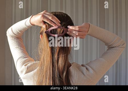 Frau, Die Langes Haar Mit Klauenklammer Stylt. Rückansicht einer Frau, die ihr langes braunes Haar mit einem rosafarbenen Krallenclip in warmem, natürlichem Innenlicht fixiert. Stockfoto