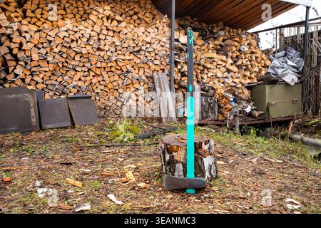 Schwere Holzspaltmühle, die in einem Baumstamm stecken und im Hintergrund einen großen Stapel Brennholz stapeln. Brennholzvorbereitung für den Winter. Stockfoto