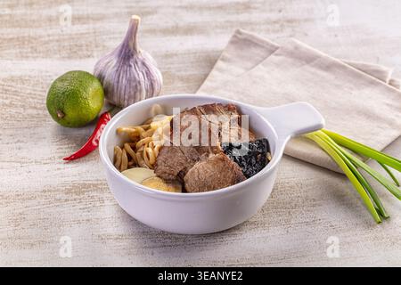 Eine köstliche Schüssel asiatischer Ramen-Nudelsuppe mit Schweinefleisch, gekochten Ei und Nori. Umgeben von frischen Zutaten wie Knoblauch, Limette und Chili auf einem rustikalen Stockfoto