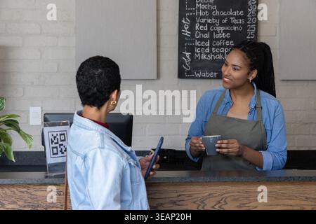 Verschiedene Kundin und Barista auf der Schürze, die am Kaffeeschalter mit Telefon und Kopierraum eine Tasse austauschen. Kaffee, Laden, Tafel, Brick, Lächeln, qr, zahlende Mitarbeiter Stockfoto