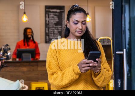 Asiatische Frau, die in der Nähe des Eingangs mit Smartphone am Café steht und einen senfgelben Pullover trägt. Kaffee, Barista, gemütlich, Ambiente, Espresso, Chalkb Stockfoto