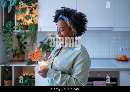 Glückliche schwarze Frau genießt die glühende Aromakerze in den Händen und fühlt Dankbarkeit, einfache tägliche Freuden zu Hause Stockfoto