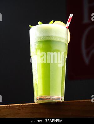 Grüner Smoothie in hohem Glas, gekühlt und schaumig. Minzzweig und Zitronenscheibe zieren den Rand. Daneben liegt ein rot-weiß gestreifter Strohhalm. Holzfläche Stockfoto