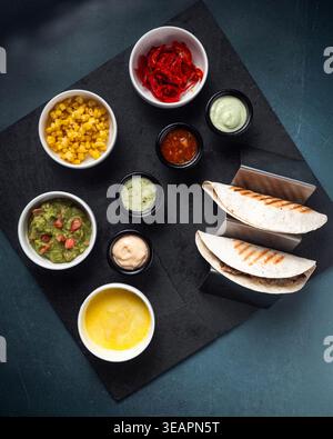 Tacos ruhen auf einem dunklen Servierbrett. Salsa, Guacamole und Beläge umgeben das Hauptgericht. Jedes Gewürz sitzt in einer eigenen kleinen Schüssel. Gegrillte Tortilla Stockfoto