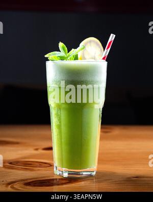 Frischer grüner Smoothie im hohen Glas mit Minze- und Zitronenscheibe. Rot-weiß gestreifter Strohhalm sorgt für verspielten Kontrast. Leicht schaumige Oberseite lässt cremige Töne vermuten Stockfoto