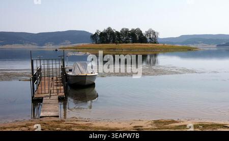 Malerischer Blick auf das Batak Reservoir mit hölzernem Pier, kleinem Boot und einer mit Kiefern bedeckten Insel, die sich im ruhigen Wasser spiegelt, Rhodope Mountains. 2023 Stockfoto