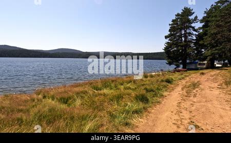 Sonniger Blick auf den See am Golyam Beglik Reservoir mit unbefestigter Straße, Kiefernwald, Campingplatz und ruhigem blauem Wasser im Spätsommer. 2023 Stockfoto