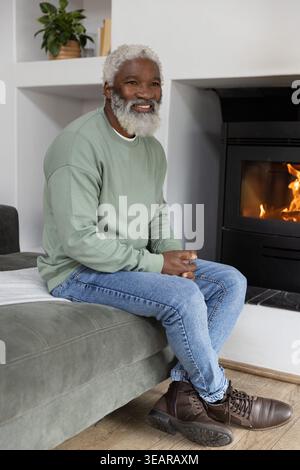 Älterer Afroamerikaner sitzt auf Sofa im Wohnzimmer und trägt ein Sweatshirt, das am Kamin lächelt. Gemütlich, Zuhause, Wärme, Herd, Feuer, Holzverbrennung, Couc Stockfoto