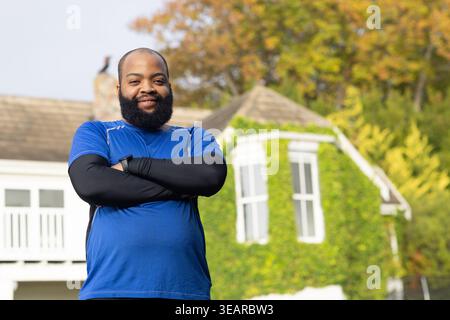 Afroamerikanischer Erwachsener, der mit gekreuzten Armen auf dem Vorstadtrasen steht und blaues Hemd trägt. Außenbereich, Wohnbereich, Innenhof, Efeu, Herbst, Bär Stockfoto