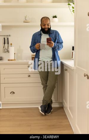 Afroamerikaner lehnt sich auf die Küchenzeile, hält einen grauen Becher, während er das rote Smartphone überprüft. Modern, gemütlich, Zuhause, Innenraum, Regale, Pflanzen, Appli Stockfoto