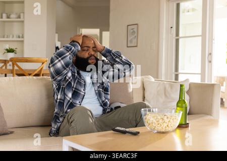 Afroamerikaner sitzt zu Hause auf beigefarbenem Sofa, hält Kopf, kariertes Hemd, Popcorn auf dem Tisch. Frustration, Stress, besinnlich, Wohnzimmer, Sunli Stockfoto