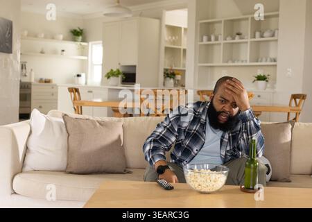 Bärtiger Mann mit kariertem Overshirt, der auf der rechten Seite des Sofas sitzt und die Fernbedienung neben der Popcorn-Schüssel hält. Offen, Zuhause, offener Plan, Wohnzimmer, Pflanzen, Flasche Stockfoto