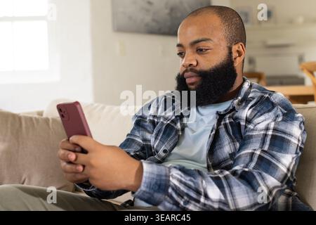 Afroamerikaner sitzt auf beigefarbenem Sofa mit rotem Smartphone, schaut auf den Bildschirm, Kopierraum. Zuhause, Innenraum, Tageslicht, Lifestyle, Ruhe, gemütliche Einrichtung Stockfoto