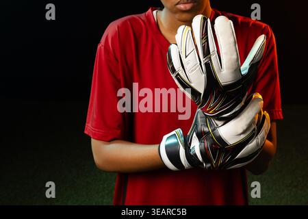 Afroamerikanerin, die Torwarthandschuhe mit Handgelenkriemen aus rotem Jersey hält und auf dem Rasen steht. Sportlich, Fokus, Intensität, dramatisch, Randbeleuchtung, Stockfoto