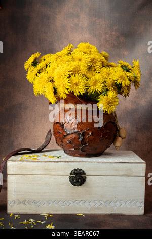 Vertikales Stillleben von gelben Löwenzahn in einem rustikalen Tontopf, der auf einer alten weißen Holzkiste liegt. Elegante Blumenkomposition in warmen Tönen. Stockfoto